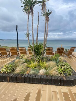 Eine Holzterrasse mit Stühlen, die Blick auf das Meer, Palmen und verschiedene Pflanzen bietet.