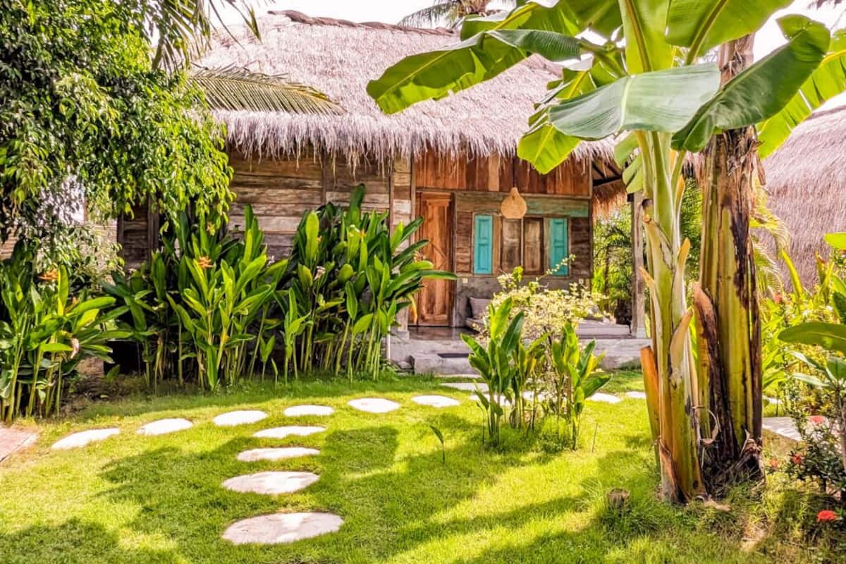 Una casa de madera rústica con tejado de paja, rodeada de abundante vegetación y un camino de piedra.