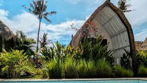 Zona de piscina con una estructura de techo de paja, exuberante vegetación y un cielo azul claro.