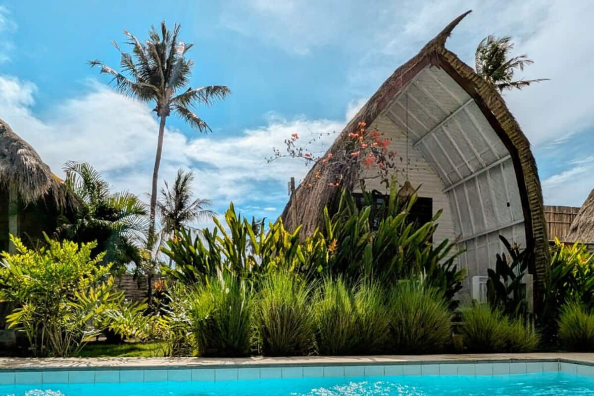 Zona de piscina con una estructura de techo de paja, exuberante vegetación y un cielo azul claro.