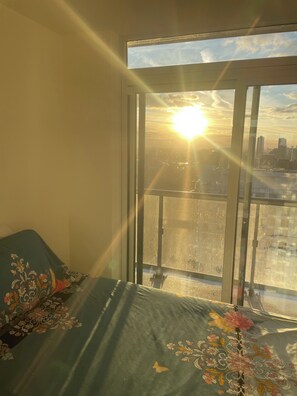 A room with a bed covered in a floral-patterned blanket, a window with a view of a city skyline, and a sun shining through the window.