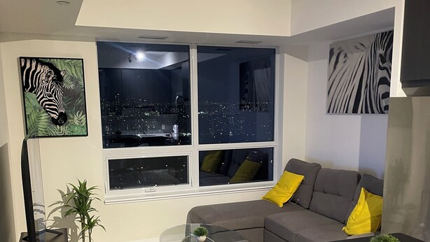 A modern living room with a city view, a sofa with yellow cushions, a glass coffee table, and a TV on the wall.