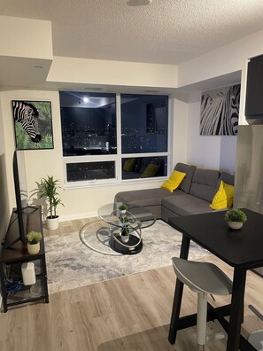 Un salon moderne avec vue sur la ville, un canapé avec des coussins jaunes, une table basse en verre et un téléviseur fixé au mur.