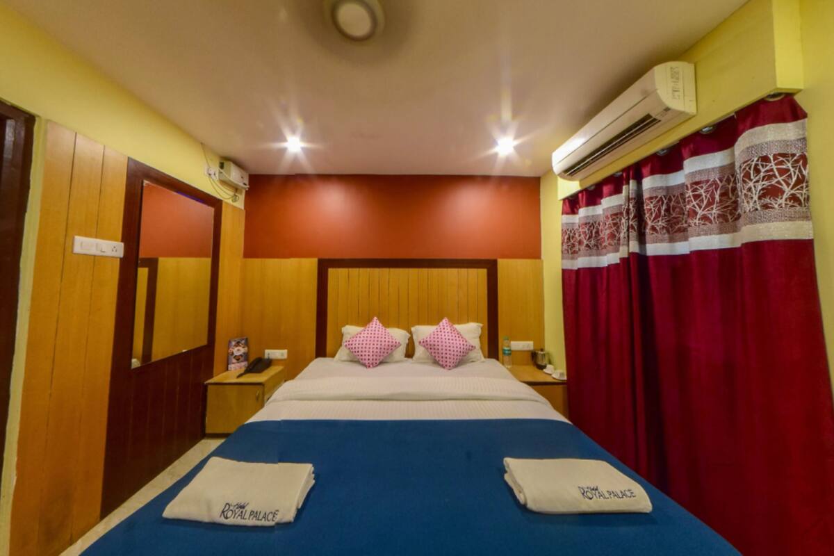 Quarto de hotel com cabeceira de madeira, cama com roupa de cama azul, duas almofadas, mesa de cabeceira com abajur e cortina vermelha.