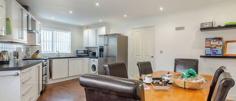 A modern kitchen with white cabinets, a built-in oven, microwave, and washing machine. There is a dining area with a wooden table and chairs.