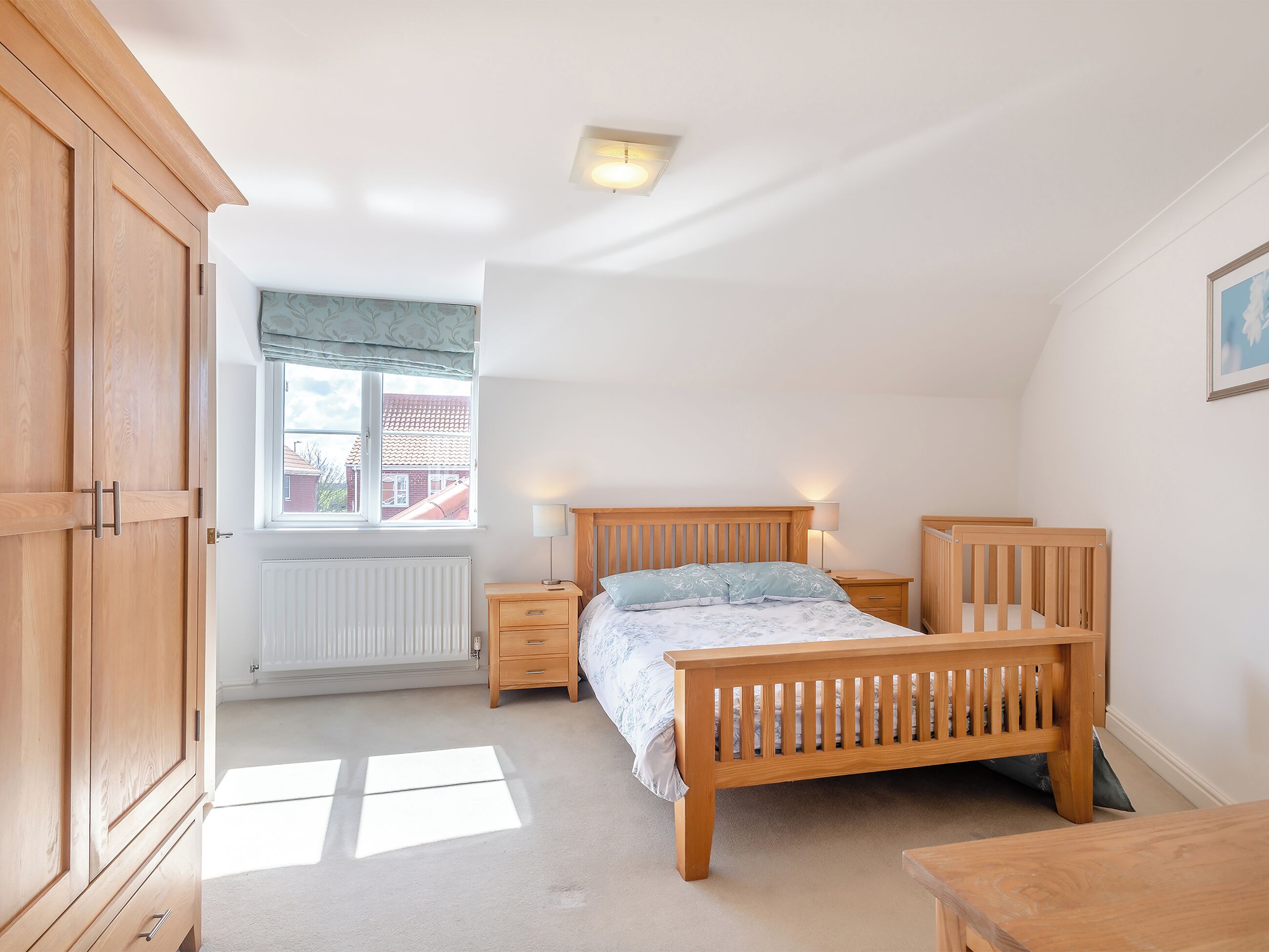 A bedroom with a wooden bed, bedside tables, a wardrobe, a radiator, and a window with a view of a house.