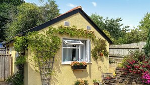 Ein kleines Haus mit geneigtem Dach, einem Fenster mit weißem Rollo und mehreren Blumentöpfen rund um die Fassade.