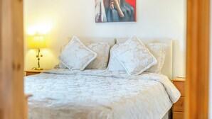Een opgemaakt bed met een bloemenprint deken en twee decoratieve kussens, een bedlamp en een schilderij aan de muur.
