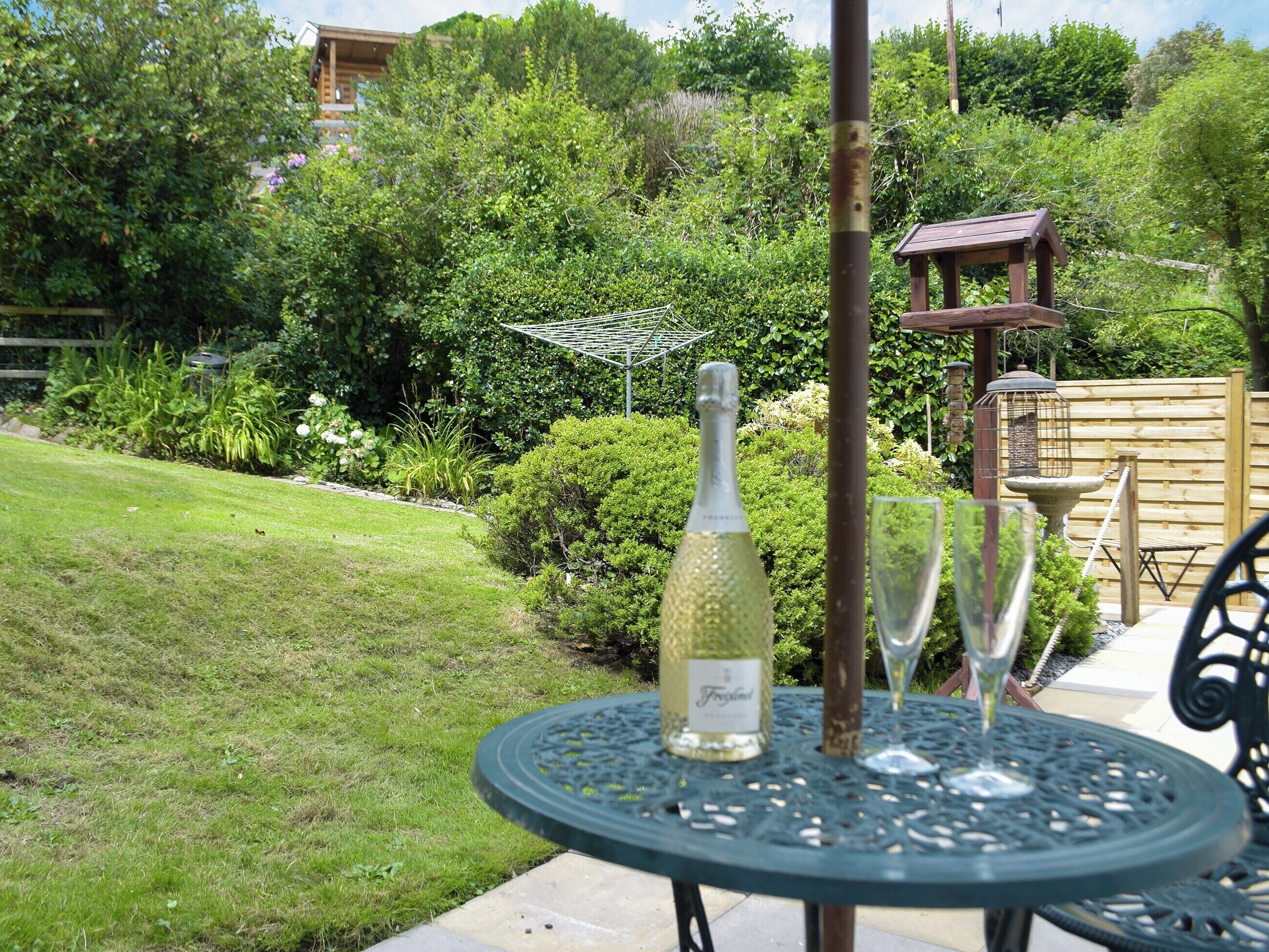 A round metal table with a bottle and two glasses on it, set in a garden with a wooden fence and greenery in the background.