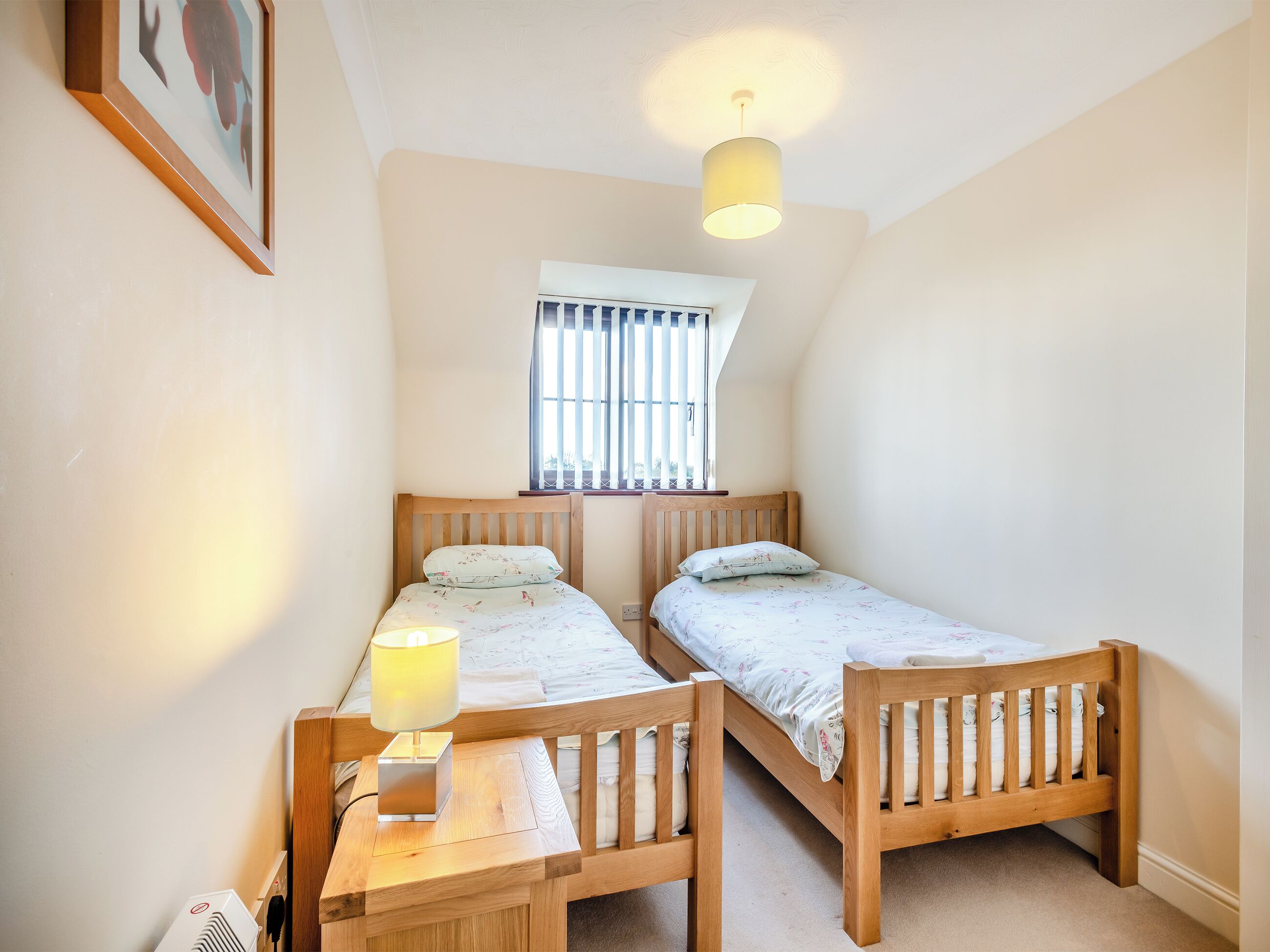 A bedroom with two wooden beds, a bedside table with a lamp, a window with blinds, and a ceiling light fixture.