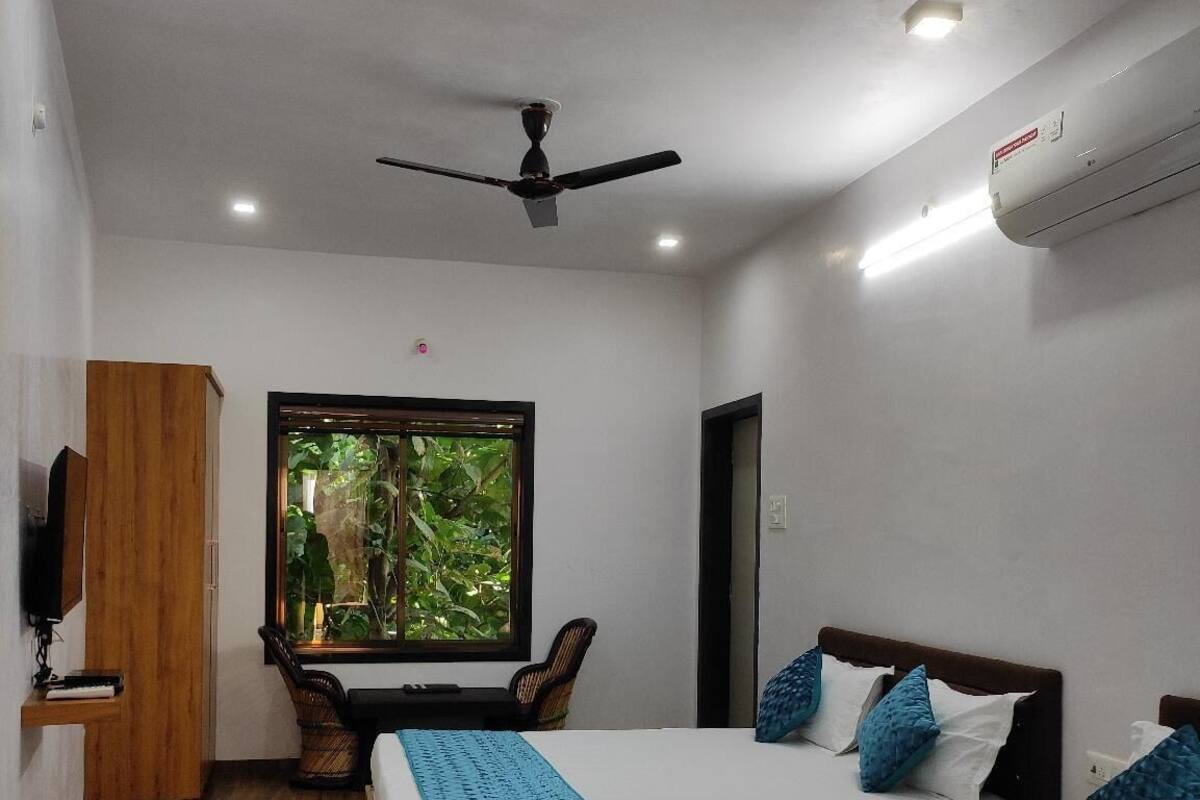 Une chambre d’hôtel avec deux lits, un ventilateur de plafond, un climatiseur, une fenêtre donnant sur un espace verdoyant et une armoire en bois.