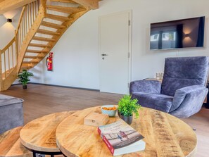 Ein modernes Wohnzimmer mit einer Holztreppe, einem grauen Sessel, einem runden Holztisch mit Büchern und einer Pflanze sowie einem an der Wand montierten Flachbildfernseher.
