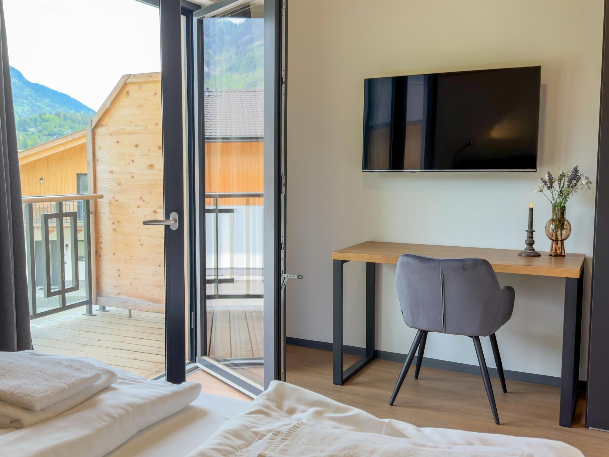 A bedroom with a bed, a desk, a chair, a TV, and a balcony with a wooden exterior.