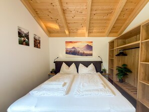 Ein Schlafzimmer mit Holzbalkendecke, einem Bett mit weißer Bettwäsche, einem Wandbild mit Berglandschaft und einem Holzschrank.