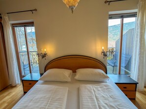 Una cama doble con dos almohadas, cabecera de madera, mesitas de noche con lámparas y ventanas amplias con cortinas.