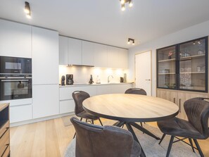 A modern kitchen with white cabinets, a built-in oven, and a dining area with a round table and leather chairs. - Apartment with sunny terrace and parking (Middelkerke)