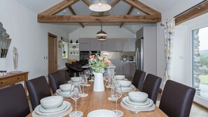 Een eetkamer met een houten tafel gedekt voor zes personen, een keuken op de achtergrond en uitzicht op het landschap door een groot raam.