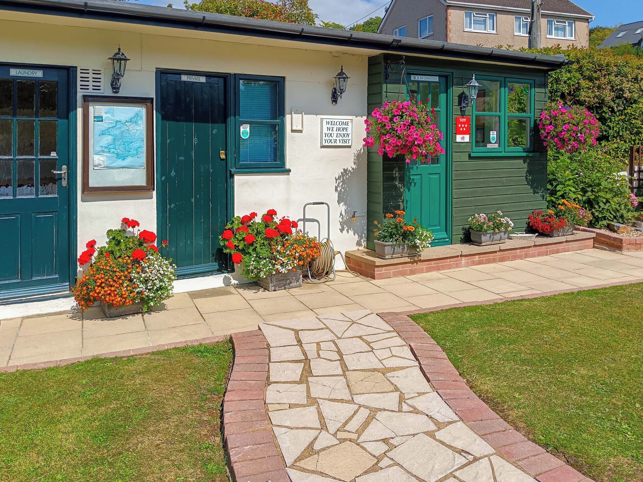 Un bâtiment avec une porte verte, une enseigne sur laquelle on peut lire « Bienvenue, nous espérons que votre visite vous plaira », et des jardinières remplies de fleurs devant l’entrée.
