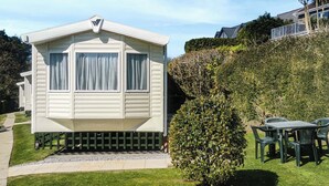 Une maison mobile de plain-pied, dotée d’un toit plat, de deux fenêtres avec des rideaux, d’un petit porche à l’avant et d’une terrasse avec une table et des chaises.