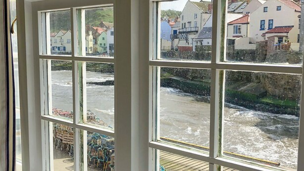 A view through a window with multiple panes, overlooking a coastal area with buildings and a dock. - 2 bedroom accommodation in Staithes, near Whitby (Staithes, near Whitby)