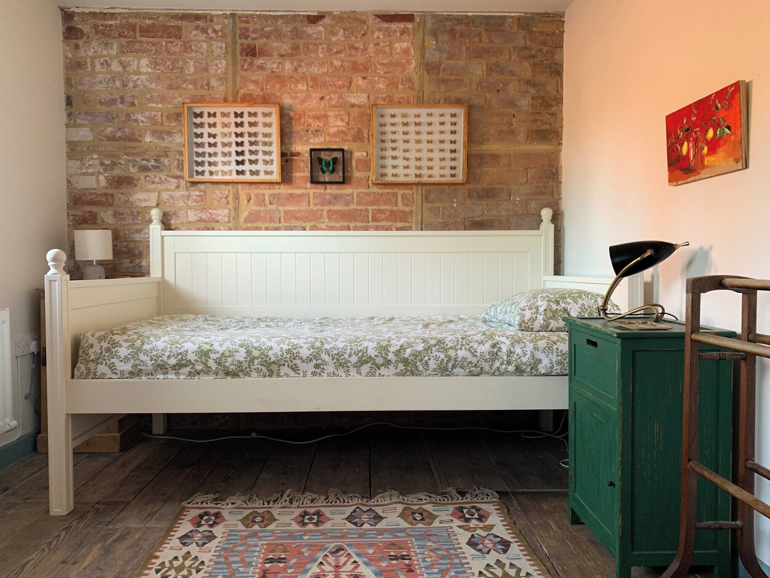 Ein Schlafzimmer mit Backsteinwand, einem weißen Bett, einer grünen Kommode und einem gemusterten Teppich.