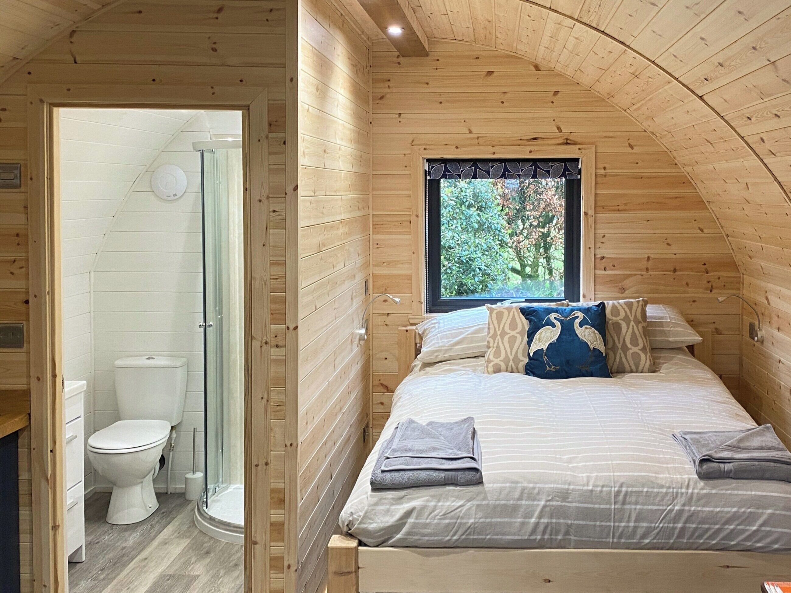 Camera da letto in una baita di legno, con un letto, cuscini e una finestra vista su un paesaggio verde.