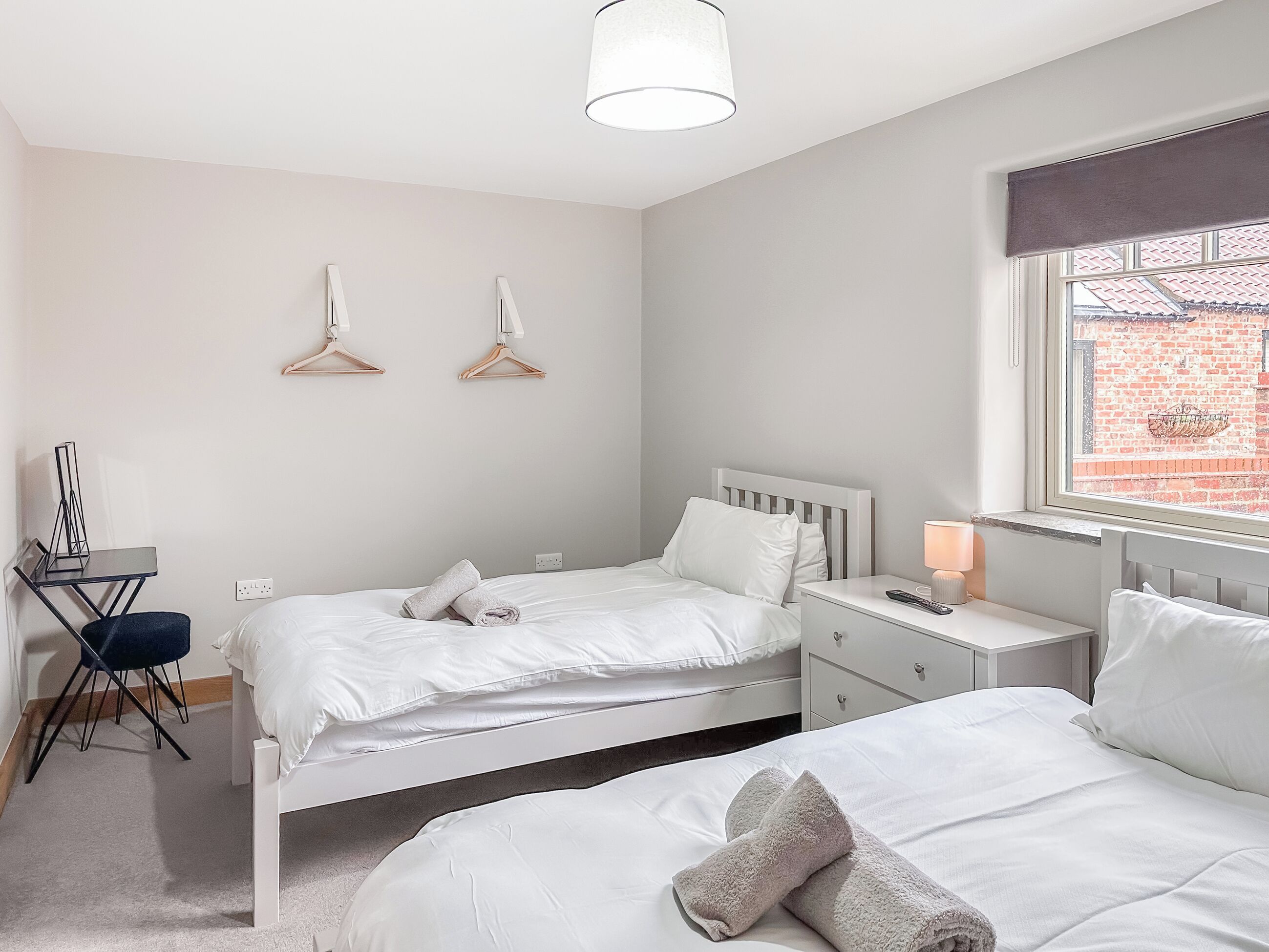 Une chambre avec deux lits, un bureau, une chaise et une fenêtre donnant sur un immeuble en briques.