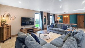 En moderne stue med et stort fladskærms-tv, grå sofaer, et sofabord, træskabe og et spiseområde med bord og stole.