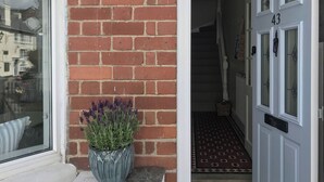 En rød murstensvæg med en potteplanter med lavendel på en hvid sokkel, en hvid dør med nummer 43, og et mønstret gulv i gangen.