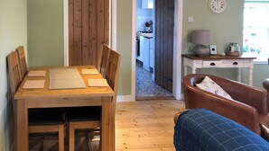 Un comedor con una mesa y sillas de madera, una cocina visible a través de una puerta abierta y un reloj en la pared.