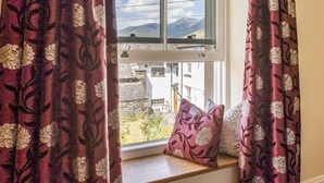 Une fenêtre ornée de rideaux à motifs floraux, une vue sur une montagne et un banc avec des coussins.