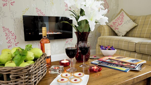 A wooden table with a bottle of wine, glasses, a candle, a basket of apples, a vase with white flowers, a plate with mini pies, and a magazine. - 2 bedroom accommodation in Keswick (Keswick)