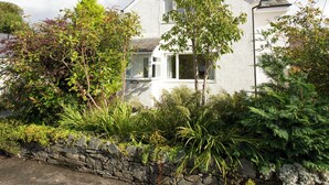A two-story house with a stone wall in front, surrounded by various plants and trees. - 2 bedroom accommodation in Keswick (Keswick)
