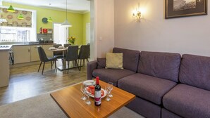 A modern living room with a purple sofa, a wooden coffee table with a bottle of wine and glasses, a dining area with a table and chairs, and a kitchen in the background. - 3 bedroom accommodation in Keswick (Keswick)
