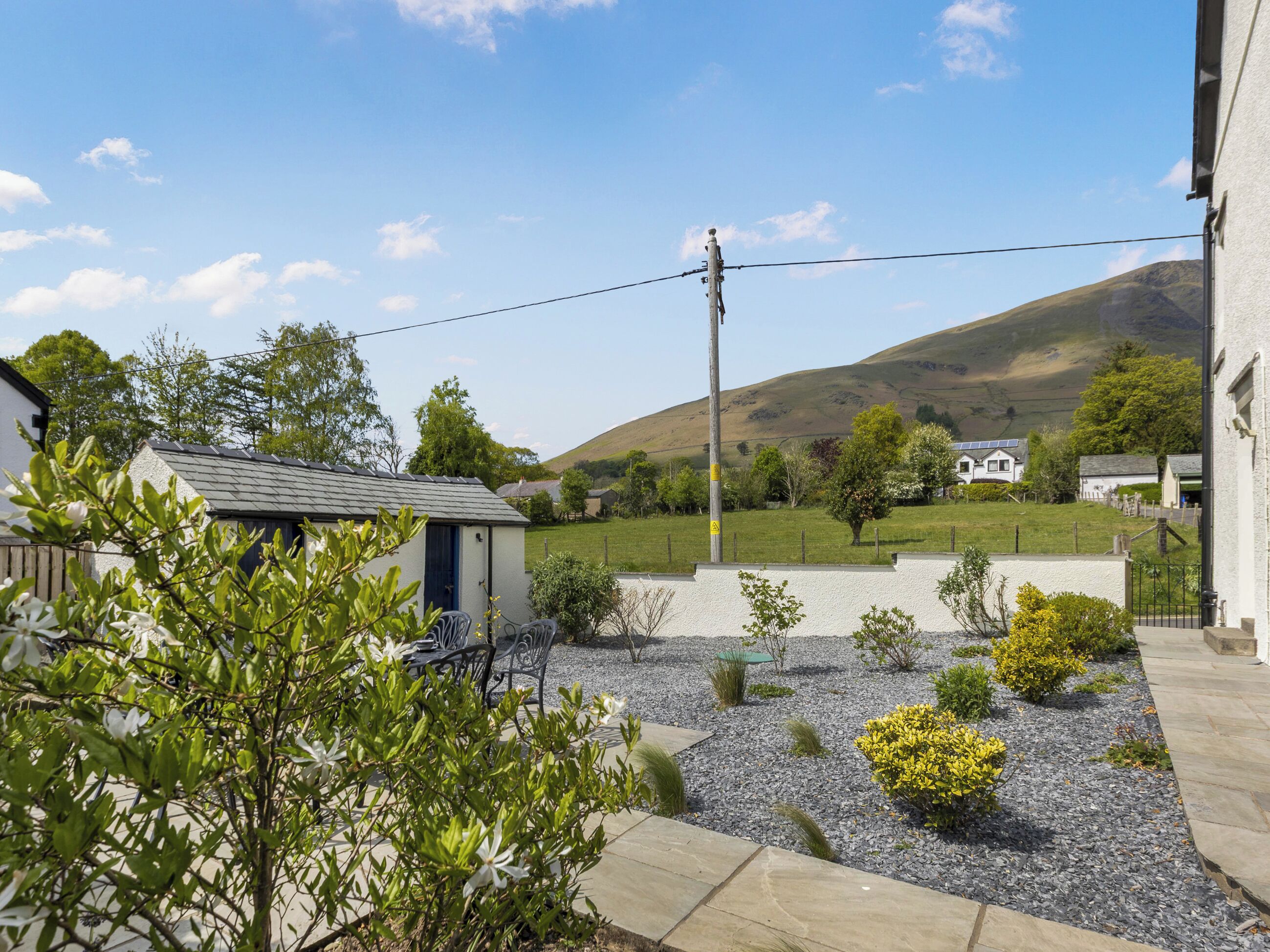 A residential area with a house, a small garden with gravel and plants, a clear sky, and a hill in the background.