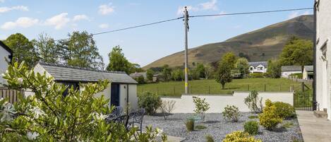 A residential area with a house, a small garden with gravel and plants, a clear sky, and a hill in the background.