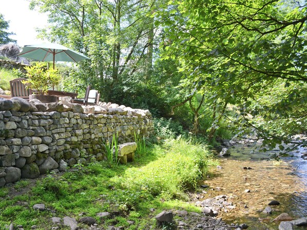 A stone wall with a chair and umbrella, a clear stream, and lush greenery. - 2 bedroom accommodation in Branthwaite, near Caldbeck (Branthwaite, near Caldbeck)