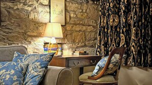 A cozy room with a stone wall, a wooden desk, a lamp, a patterned chair, and a blue floral sofa. - 2 bedroom accommodation in Pooley Bridge (Pooley Bridge)