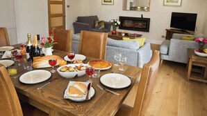 Ein Essbereich mit Holztisch, gedeckt für eine Mahlzeit, ein Wohnzimmer mit Sofa und Fernseher sowie eine Küche im Hintergrund.