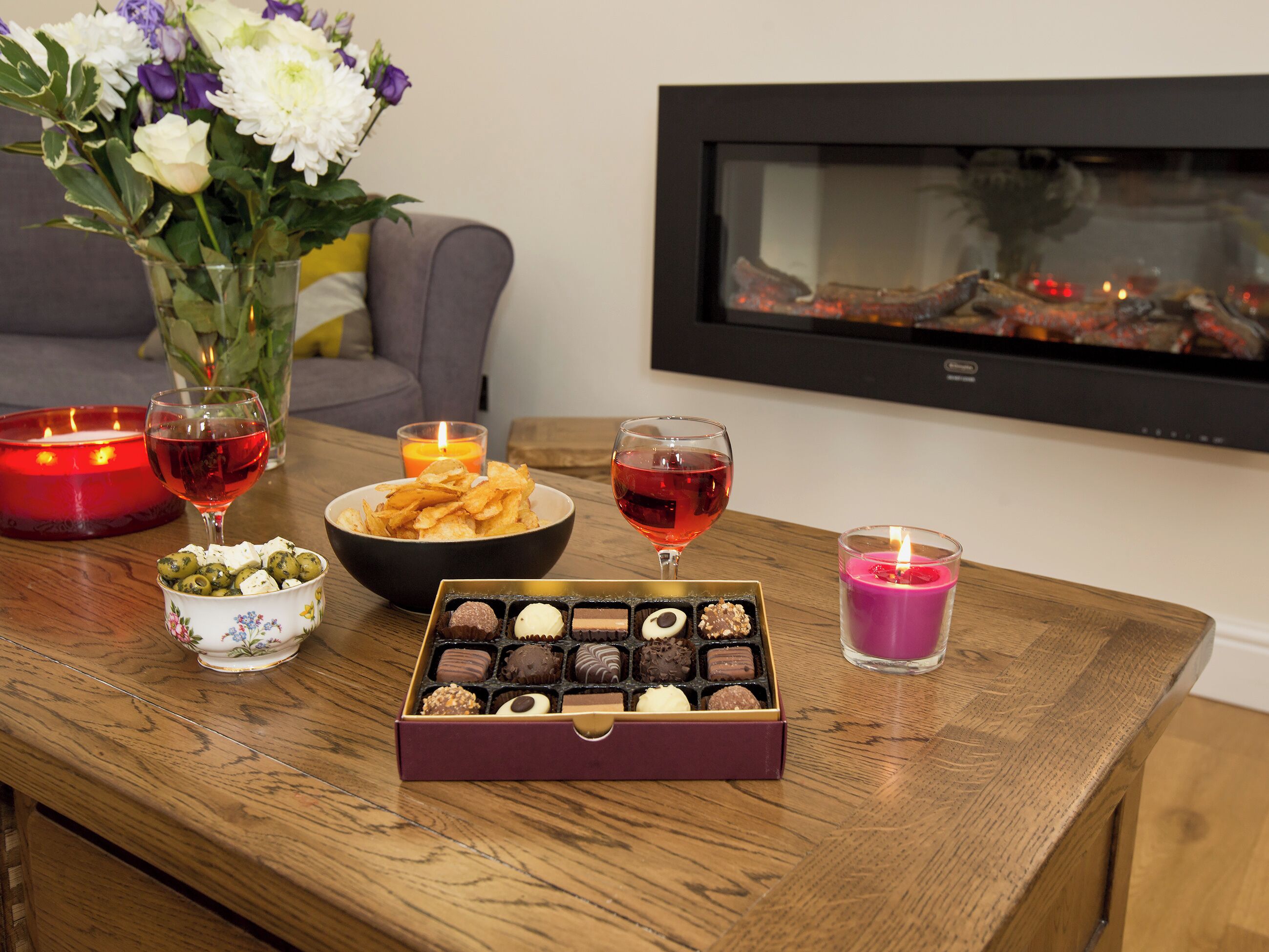 A wooden table with a box of chocolates, a bowl of chips, a glass of red wine, a candle, and a vase of flowers.