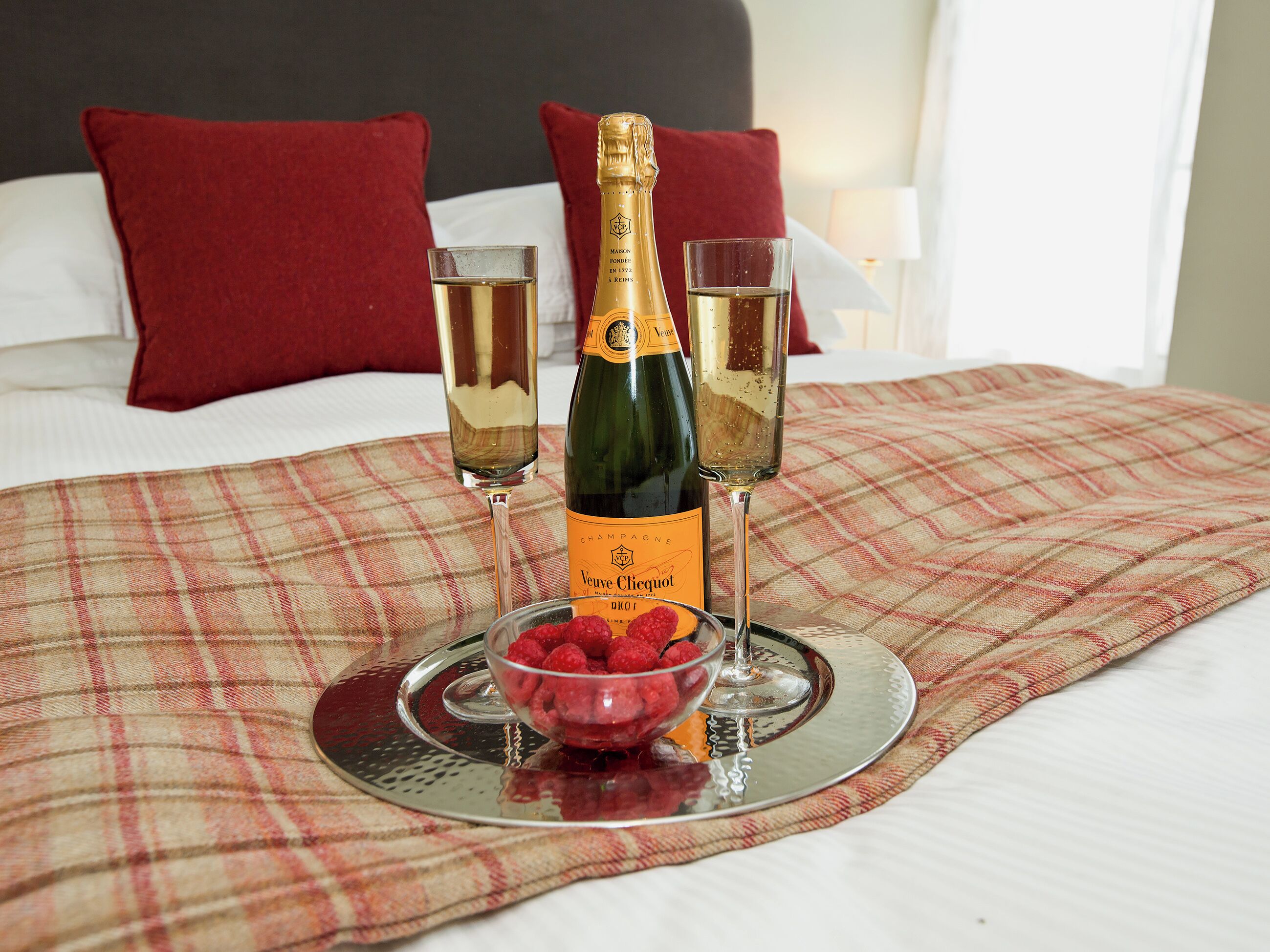 A bed with red pillows, a bottle of Veuve Clicquot champagne, two glasses of champagne, and a bowl of raspberries on a plaid blanket.