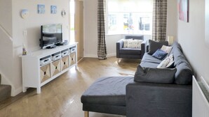 En stue med en grå sofa, et hvidt tv-møbel, et fjernsyn og et vindue med gardiner.