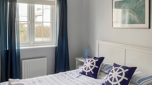 Et soveværelse med en seng, to puder med et skibsrod design, et vindue med gardiner, en radiator og et billede i ramme på væggen.