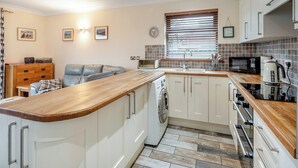 A compact kitchen with a wooden countertop, white cabinets, a built-in washing machine, and a window with blinds. - 1 bedroom accommodation in Red Row near Amble (Red Row near Amble)