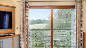A room with a TV, a stone wall, and a glass door leading to an outdoor area with chairs. - 1 bedroom accommodation in Red Row near Amble (Red Row near Amble)