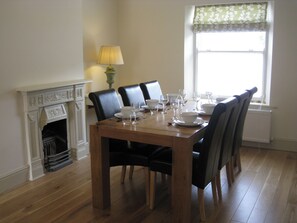 A dining room with a wooden table set for six, black chairs, a white fireplace, and a window with floral curtains. - 3 bedroom accommodation in Bradwell (Bradwell)