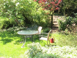 Een tuin met een ronde tafel en twee stoelen, omgeven door weelderige begroeiing en een kleine rode emmer.