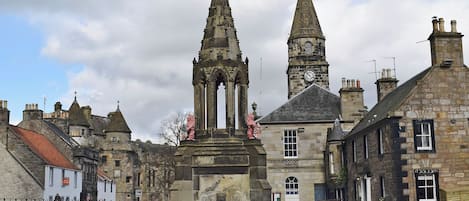 Uma praça histórica com uma torre do relógio imponente, edifícios de pedra e um monumento central.