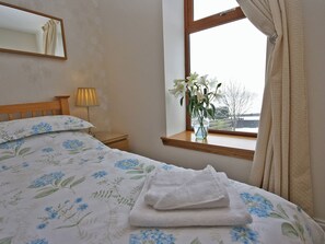 Una cama bien hecha con ropa de cama floral, una lámpara de noche, un florero con flores en el alféizar de la ventana y una ventana con cortinas.
