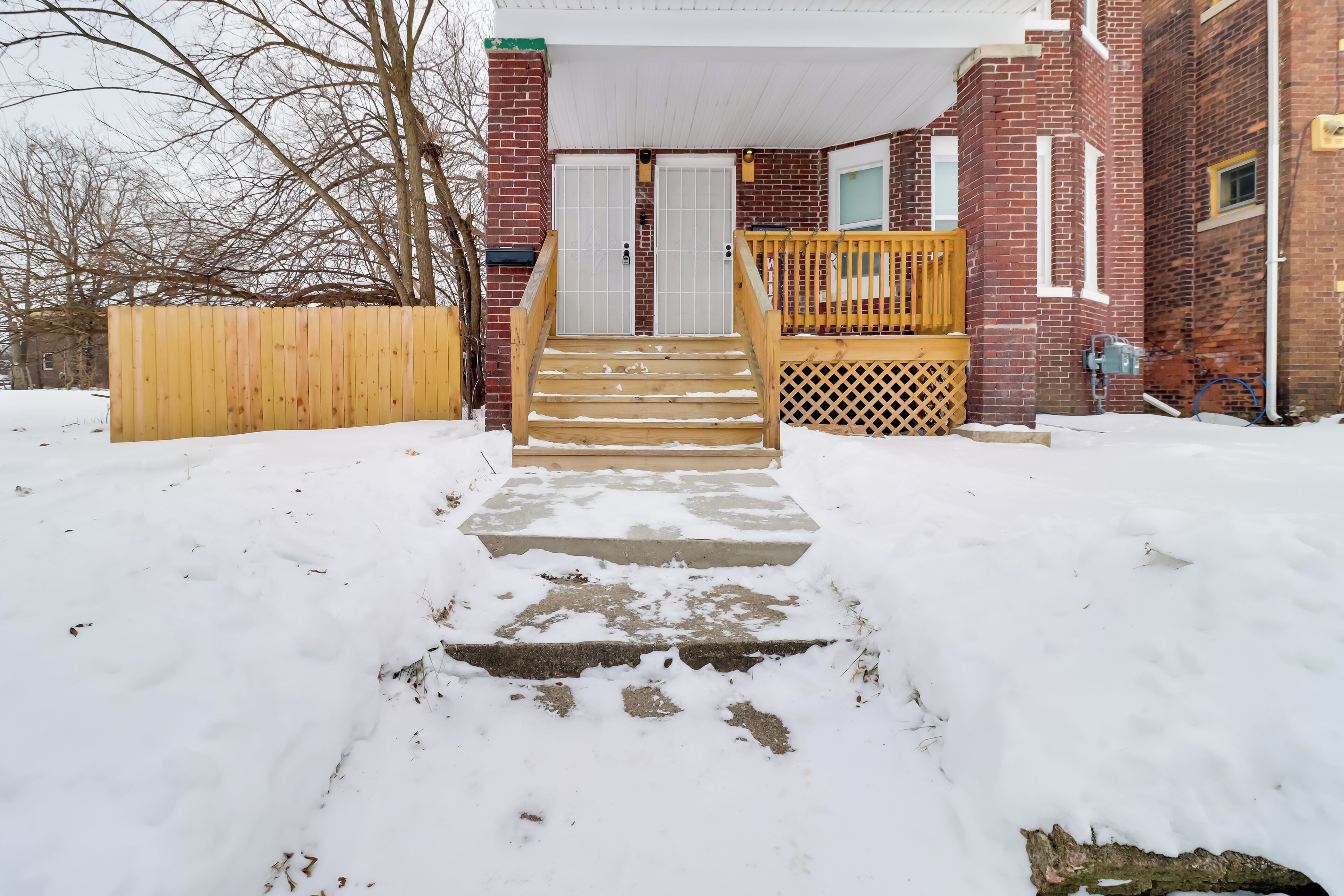 A snow-covered front yard leading to a brick house with a wooden deck and a front door.